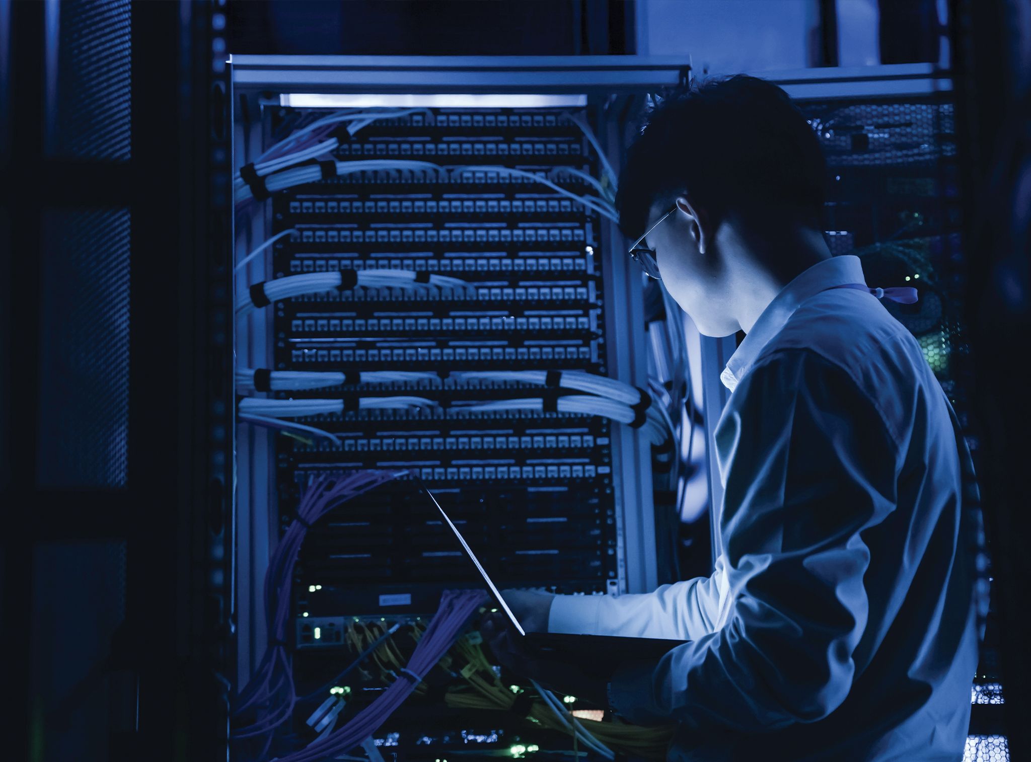 A technician holding a laptop inspects a server rack with bundled network cables in a dimly lit, blue-toned data center